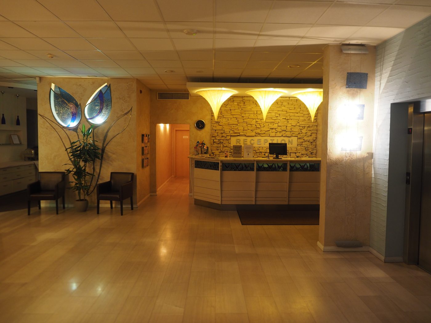 An empty hotel reception desk late at night, lit and tidy, with a monitor on the counter, a sign reading RECEPTION on the stone wall behind it, and nobody there