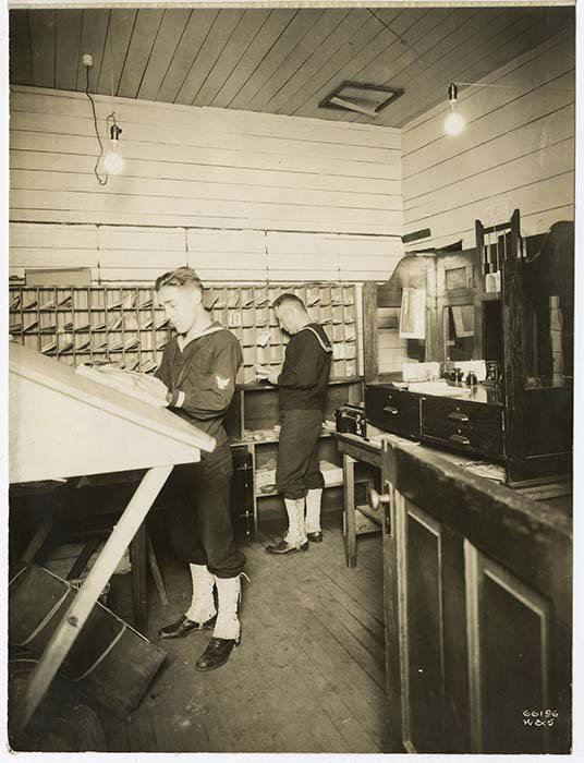 Two sailors in a wood-paneled mail room around 1918, one reading letters at a slanted desk, the other at a wall of pigeonhole sorting boxes
